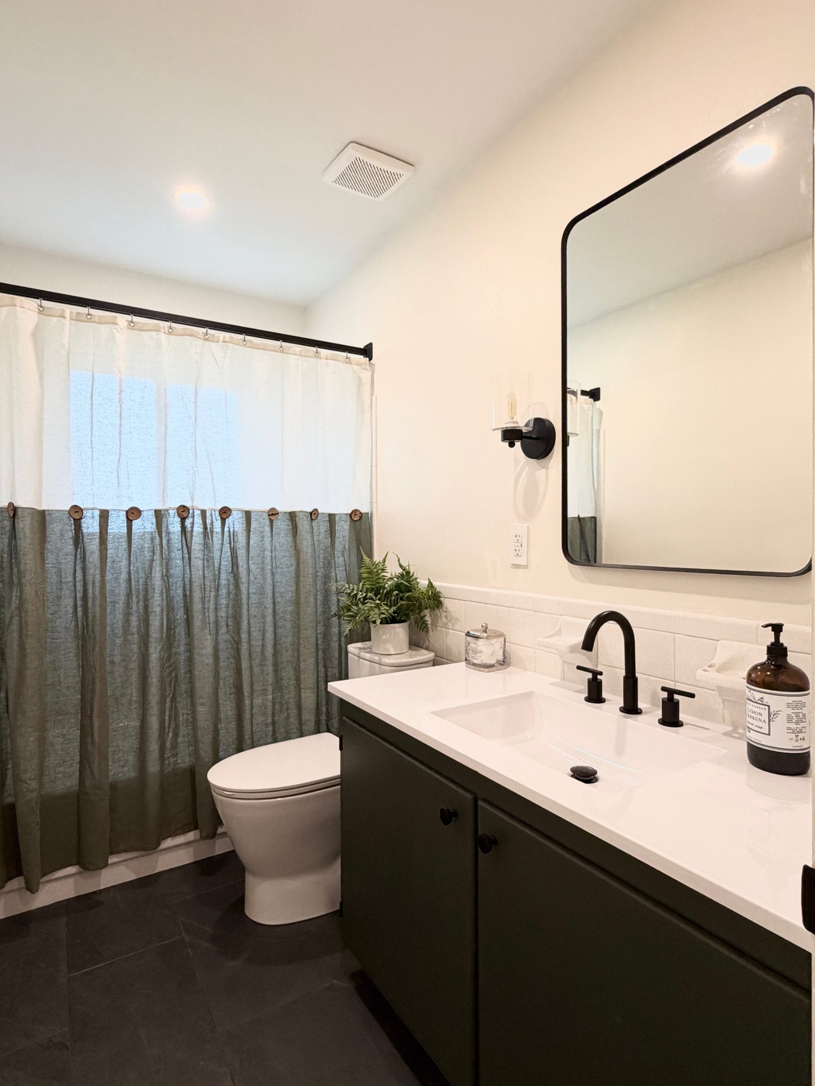 Finished hall bath with a deep green vanity, white top, black fixtures, a rounded mirror and a green linen shower curtain