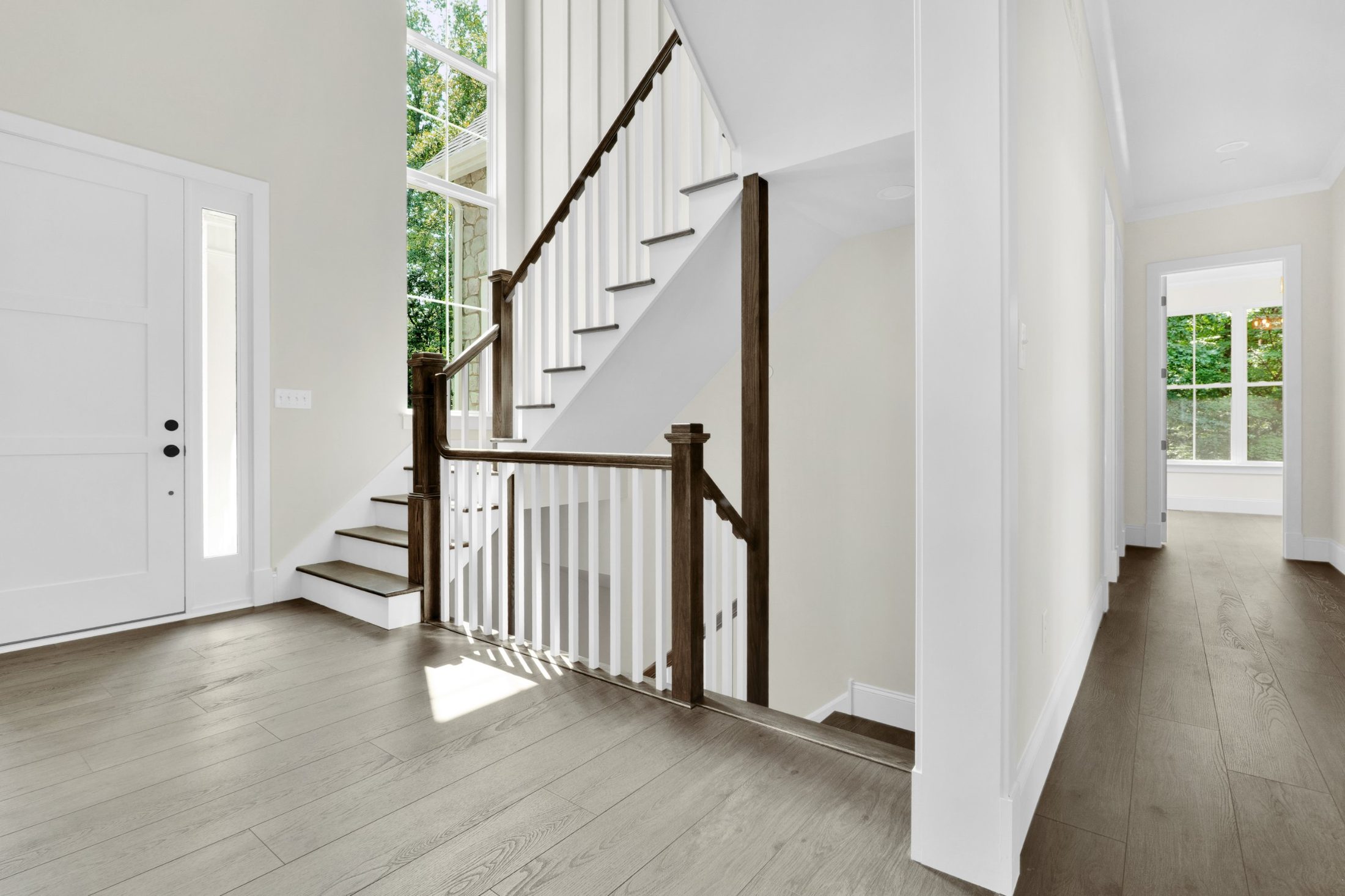 Entry foyer with a white front door, open staircase with dark wood rail and white balusters, and a hall leading to a bright room