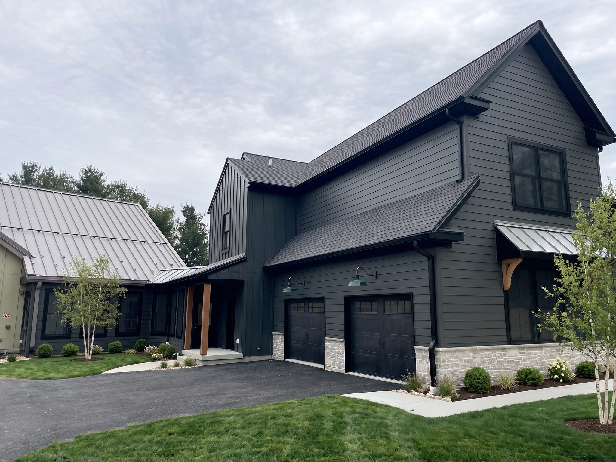 Front of the home: charcoal siding and board-and-batten, two-car garage with green barn lights, stone base, standing-seam metal awning with cedar bracket, and a cedar-post entry beside the sage-green wing
