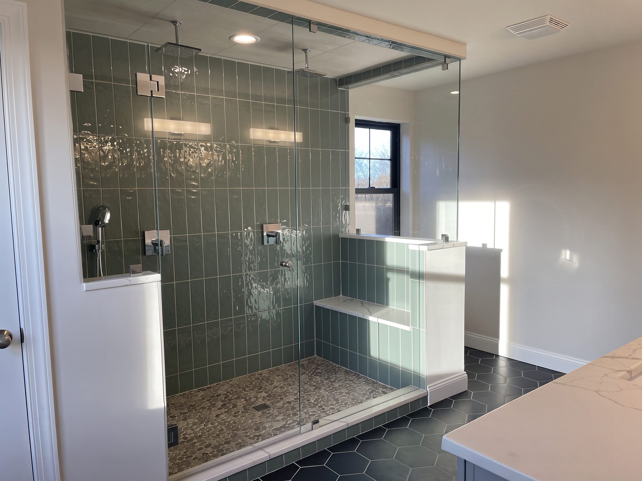 Glass-enclosed shower with green vertical tile, dual rain heads, built-in bench and pebble floor