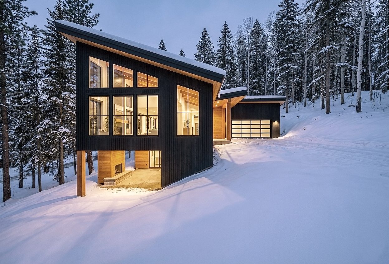 Modern home with dark vertical siding, a sloped roof and large lit windows in a snowy evergreen forest