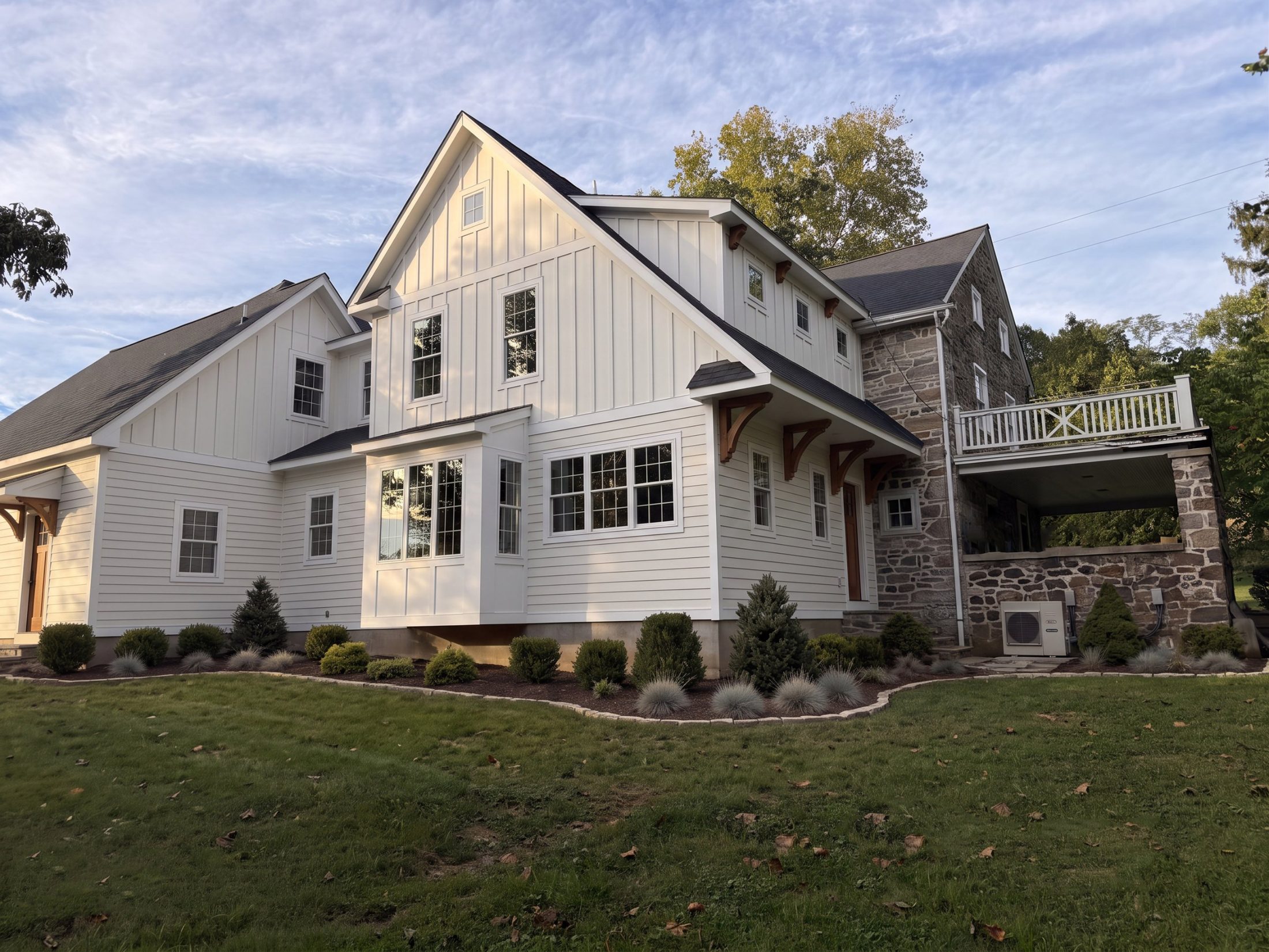 The stone farmhouse after the addition, seen from the side: white board-and-batten gable, cedar brackets, bay window and a new deck over the stone porch