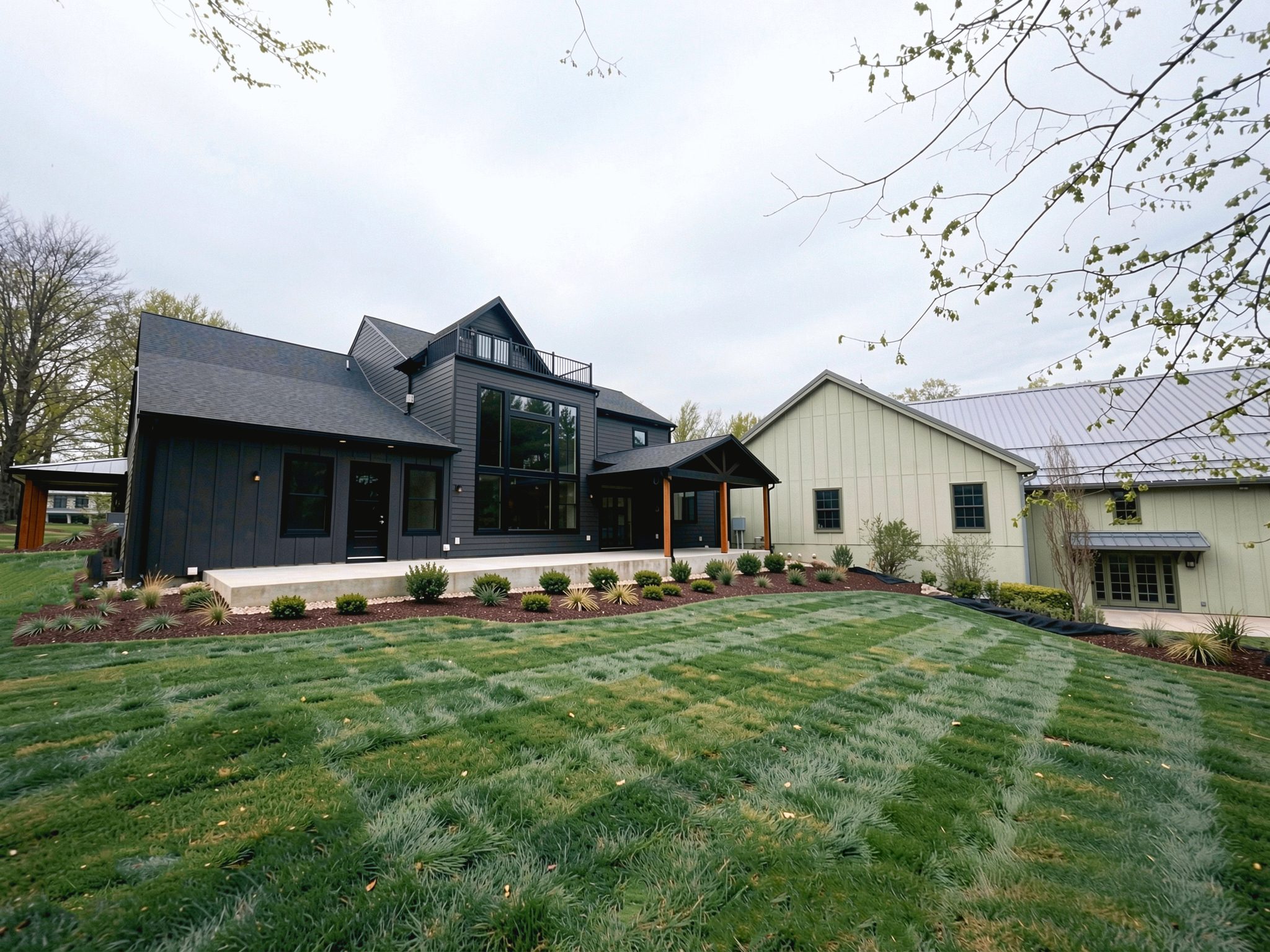 Rear of the home under an overcast sky: dark board-and-batten wing with a tall glass bay, timber-framed entry, sage-green wing with metal roof, concrete patio and new landscaping